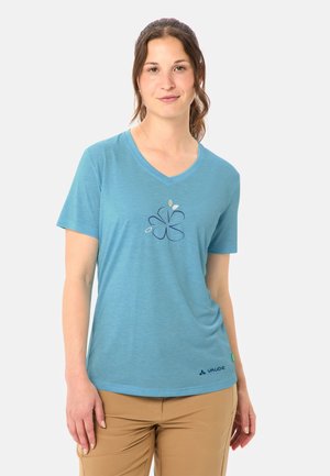 Junge Frau mit zurückgebundenem Haar, die ein blaues V-Ausschnitt-T-Shirt mit floralem Muster und beige Hosen trägt, stehend vor einem weißen Hintergrund.