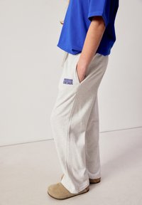 Isik, kes kannab heledaid hallid dressipükse, millel on "American Vintage" logo, sinist särki ja beeže libisevaid kingi, seisab ühe käega taskus.