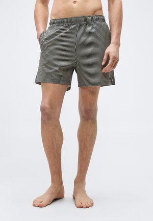 Mand iført sort-hvide shorts med lodrette striber og elastisk talje, stående barfodet mod en ensfarvet baggrund.