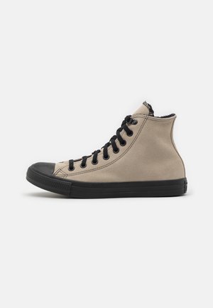 Beige højtop lærredssneaker med sort gummisål, sort tåhætte og sorte snørebånd på hvid baggrund.