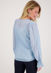 Lichtblauwe blouse met verticale strepen en een doorschijnende achterkant. Beschikt over lange puffmouwen met elastische manchetten en subtiele glinsterende accenten.