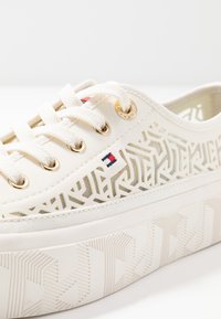 Sneaker bianca con motivi ritagliati geometrici, occhielli dorati, suola spessa e testurizzata, lacci bianchi e un piccolo logo con bandiera rossa, bianca e blu sul lato.