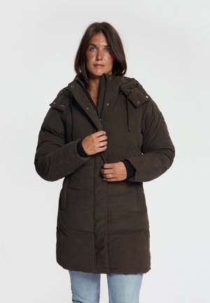Vrouw die een donkerbruine gewatteerde winterjas met hoge kraag draagt, houdt het ritsgedeelte vast, staand tegen een effen lichte achtergrond.