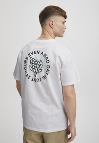 Hvid bomulds T-shirt med korte ærmer, med et grønt blomstermotiv og tekst på ryggen, designet med en afslappet pasform.