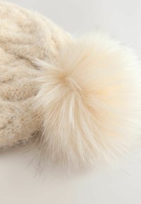 Knit cream beanie featuring a fluffy light beige pom-pom. The texture is soft with a cable knit pattern on the main body.