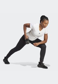 Mallas deportivas negras y una camiseta blanca de cuello en v de adidas; sosteniendo una kettlebell, ubicada en un ejercicio de estocada lateral, con una forma enfocada.