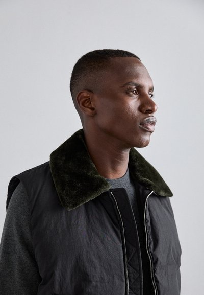 Črn brezrokavnik z zelenim umetnim krznenim ovratnikom, ki ima zadrgo spredaj in prešito podlogo, postavljen na svetlem ozadju.