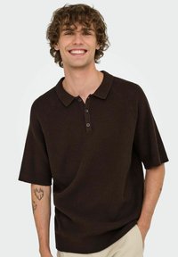 Polo de manga corta de color marrón oscuro con un tejido texturizado, cuello clásico y tapeta de tres botones. Presenta mangas raglán y un ajuste relajado.