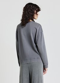 Donna con capelli ricci che guarda di lato e di spalle, indossa un ampio felpa grigia e pantaloni grigi coordinati su uno sfondo semplice.