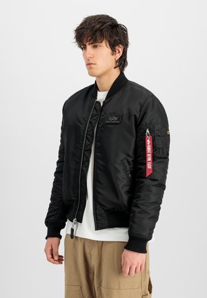 Joven con cabello oscuro despeinado que lleva una chaqueta bomber negra con una etiqueta roja "Remove Before Flight" en la manga izquierda y pantalones beige.