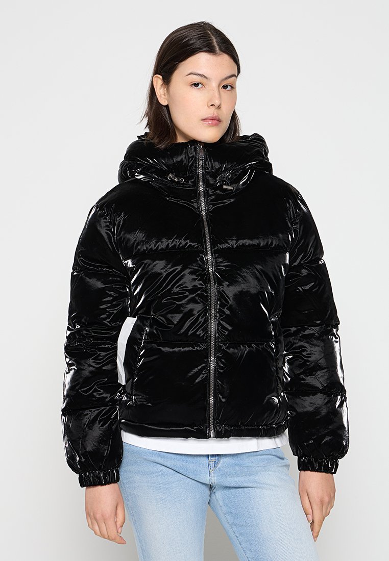 Veste doudoune noire avec finition brillante, fermeture éclair à l'avant, capuche amovible, poches latérales et manches élastiques. Portée par-dessus un haut blanc et un jean clair.