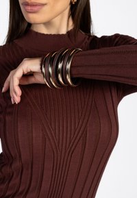 Bruine geribbelde trui met een gestructureerd verticaal patroon, voorzien van meerdere metalen armbanden in goud en bruin, gestapeld om de pols.