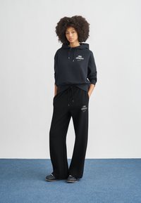 Μαύρο φούτερ με κουκούλα και γκρι λογότυπο Nike, σε συνδυασμό με μαύρο παντελόνι φόρμας με λογότυπο. Ομαλό ύφασμα, άνετη εφαρμογή και μέση με κορδόνι.