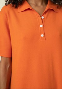 Femme portant un polo orange à manches courtes avec trois boutons blancs et un col, montrant une partie de son cou et de son bras.