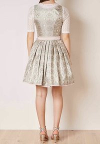 Vrouw in een geplooide bloemenjurk met kanten korte mouwen en zilveren high-heeled sandalen, staande op een houten vloer.