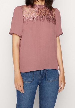 Vrouw draagt een mauve kortärmelig blouse met bloemen kant inzetstuk en halslijn detail, gecombineerd met blauwe spijkerbroek.