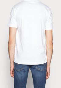 Una persona che indossa una semplice maglietta bianca e jeans blu è vista di spalle, mostrando il retro dell'outfit. Lo sfondo è di colore chiaro.