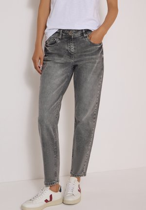 Person trägt ausgewaschene schwarze Jeans mit geradem Bein, weißes T-Shirt und weiße Sneaker mit roten und schwarzen Akzenten und steht vor einem schlichten Hintergrund.