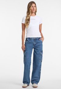 Leichtes weißes T-Shirt mit "GUESS JEANS" Logo, kombiniert mit weit geschnittenen blauen Denim-Cargohosen mit Seitentaschen und entspannter Passform.