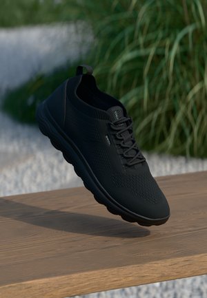 Schwarzer Sportschuh mit einem atmungsaktiven Mesh-Obermaterial, strukturiertem Design, Schnürvorderseite und gepolsteter Sohle, auf einer Holzoberfläche präsentiert.