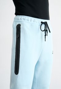 Light blue sweatpants with black zippered pockets, elastic waistband, and drawstring. Soft fabric with a relaxed fit, featuring side stitching.