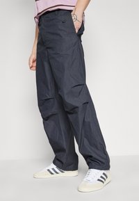 Dunkelgraue, leichte, lässig geschnittene Hose mit strukturierter Oberfläche, ausgestattet mit einem Knopfverschluss und Seitentaschen. Getragen mit weißen Sneakers.