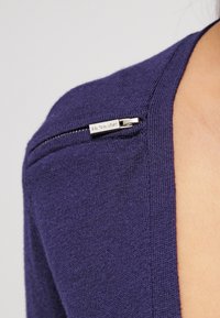 Prenda de punto morada con un detalle de cremallera en el hombro, con un tirador plateado marcado con "Morgan". Textura suave y forma ajustada.