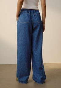 Modré džínsové nohavice so širokými nohavicami a vysokým pásom, s dvoma zadnými vreckami a jemnou textúrou, navrhnuté pre pohodlný strih.
