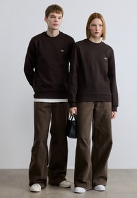 Σκούρα καφέ φούτερ με μικρά πράσινα λογότυπα, σε συνδυασμό με παντελόνια φαρδιάς γραμμής σε ανοιχτό και σκούρο καφέ. Ένα άτομο κρατά μια μικρή μαύρη τσάντα χειρός.