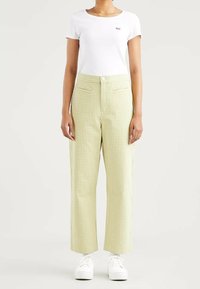 Pantalon à carreaux verts et blancs avec une taille haute, des jambes droites, deux poches avant et une fermeture à boutons, associé à un t-shirt blanc.