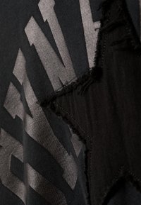 Dunkles Gewebe mit einem auffälligen, abgenutzten Sternausschnitt, ergänzt durch ein metallic graues Grafikdesign mit geometrischen Mustern.