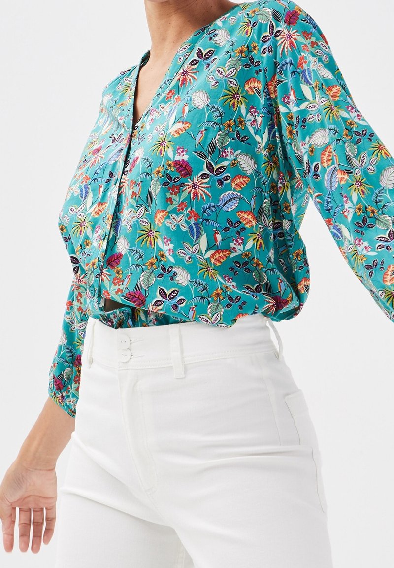 Femme portant un chemisier bleu sarcelle à imprimé floral coloré rentré dans un pantalon blanc taille haute, sur fond blanc.