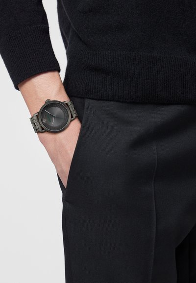 Persoon met een geheel zwarte outfit en een metalen polshorloge met een donkere wijzerplaat en een gedetailleerde rand, hand ontspannen in zak.