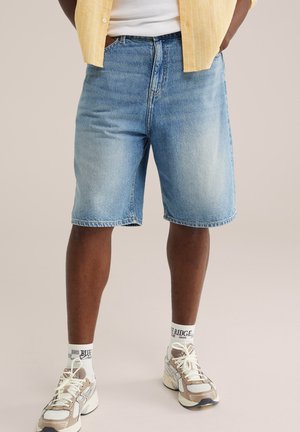 Mand iført lyseblå denimshorts, hvide sneakers, hvide sokker med sort tekst, hvid t-shirt og en gulstribet skjorte, der er åben.