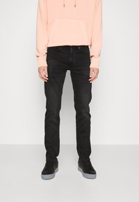 Muž v černých slim-fit džínách, černých teniskách se šedými podrážkami a světle broskvovou mikině stojí na bílém pozadí.