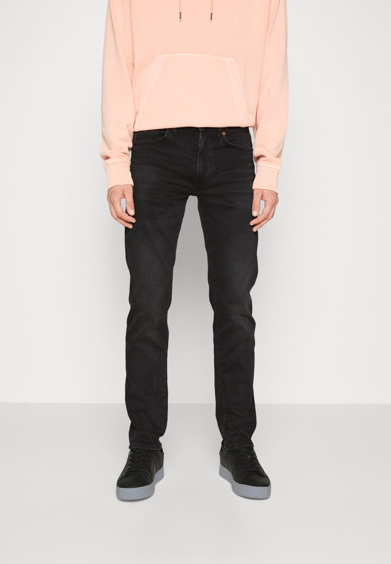 Muž v černých slim-fit džínách, černých teniskách se šedými podrážkami a světle broskvovou mikině stojí na bílém pozadí.