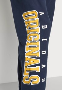 Marinblå sweatpants med fet gul och vit text "ADIDAS ORIGINALS" på vänster ben. Mjukt, avslappnat tyg med avsmalnande design.