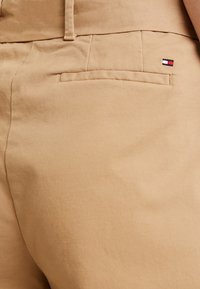 Khaki broek met een soepele textuur, voorzien van een achterzak en een klein geborduurd logo in rood, wit en blauw aan de rechterkant.