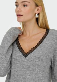 Suéter gris de punto con un escote en forma de V y delicados detalles de encaje negro. Su textura suave y las mangas largas completan el diseño.