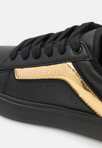 Černé tenisky s hladkým koženým svrškem, zdobené lesklým zlatým akcentem a perforovanými detaily, šněrovací s černými tkaničkami.