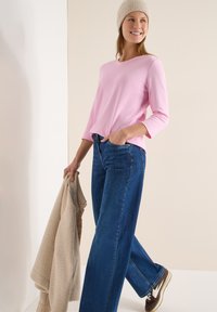Haut rose à manches longues associé à un jean large en denim bleu. Pull beige tenu dans la main gauche, baskets marron avec des accents blancs aux pieds.