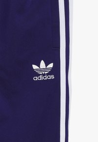 Marinblå träningsbyxor med vita sidremsor och en liten broderad Adidas-logotyp i silver på vänster lår. Slät tygtextur.