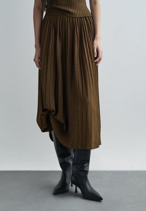 Jupe plissée marron avec un motif texturé, portée avec des bottes montantes noires à talons aiguilles, mettant en avant un mélange de matériaux lisses et côtelés.