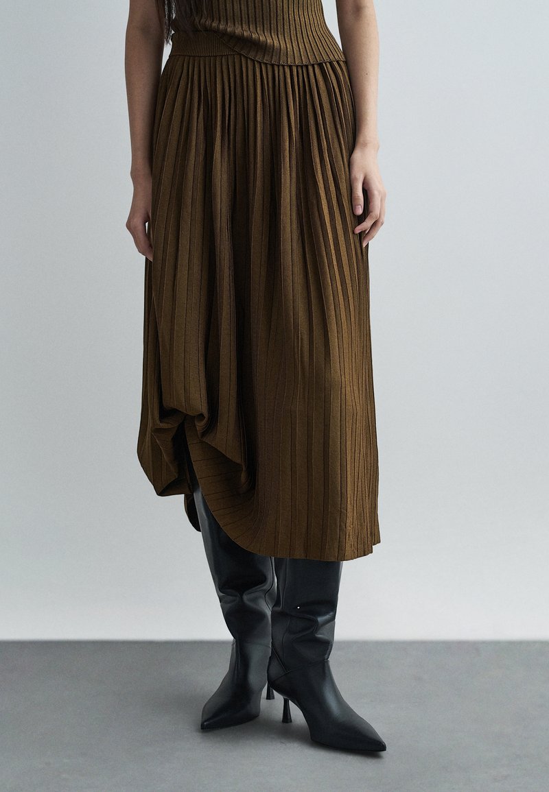 Καφέ πτυχωτή φούστα με υφή, φορεμένη με μαύρες κν�ήμες στιλέτο μπότες, αναδεικνύοντας μια μίξη λείων και ραβδωτών υλικών.