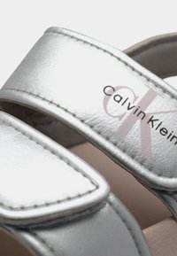 Φορεμάτα ασημί δερμάτινα σανδάλια με εμφανή ραφή και εκτυπωμένο το λογότυπο Calvin Klein σε ένα από τα λουριά.