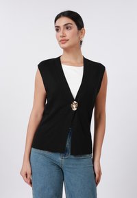 Μαύρο πλεκτό γιλέκο με βαθύ V-λαιμόκοψη και κοντά μανίκια, με χρυσό κλείσιμο σε σχήμα κεφαλής λιονταριού. Φοράται πάνω από λευκό τοπ και συνδυάζεται με μπλε τζιν.