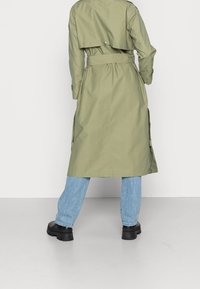 Lyse olivengrøn trenchcoat med bælte i taljen, strukturerede skuldre og flapdetalje bagpå, parret med lyseblå jeans og kraftige sorte støvler.