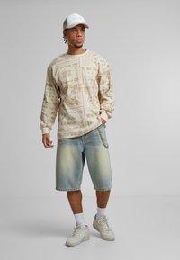 Light beige sweatshirt with intricate tan patterns, paired with faded blue denim shorts featuring a chain detail. Grey sneakers and white socks.
