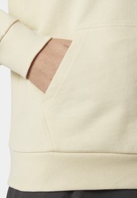 Sudadera color crema con un bolsillo frontal, puños y dobladillo acanalados. El material presenta un acabado de punto texturizado. Elementos de diseño minimalistas.