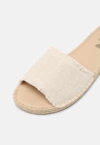 Sandale slide beige à bout ouvert avec bride en tissu texturé et détail de coutures sur la semelle, sur un fond clair.